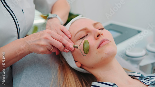 Close-up shot of facial massage with a jade roller on woman's face at beauty salon. Cosmetologist doing procedure for young woman.
