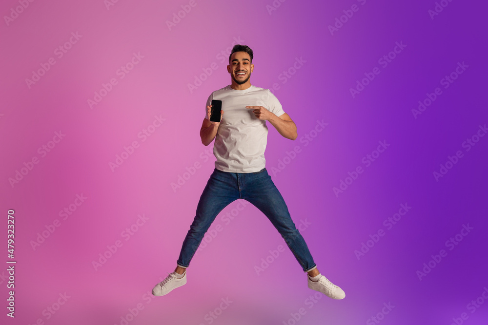 Handsome young Arab guy jumping and showing smartphone with blank screen in neon light, mockup