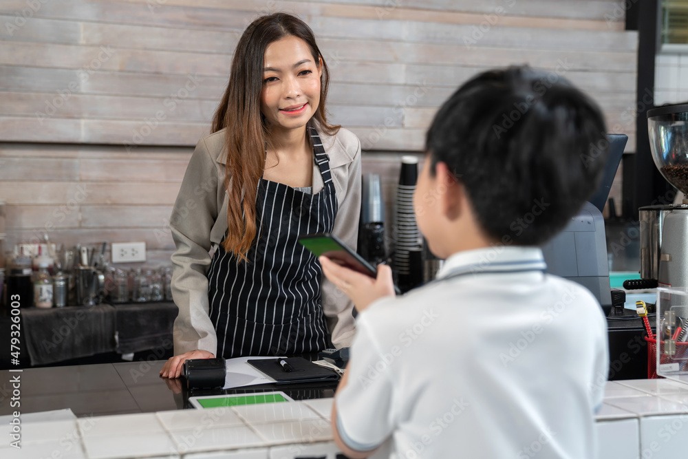 Young Asia boy order ice chocolate with barista at the coffee shop. Small business owner. Woman wearing apron looking at kid and smile. Boy using smartphone to pay drink. Technology and service. Young Asia boy order ice chocolate with barista at the coffee shop. Small business owner. Woman wearing apron looking at kid and smile. Boy using smartphone to pay drink. Technology and service.