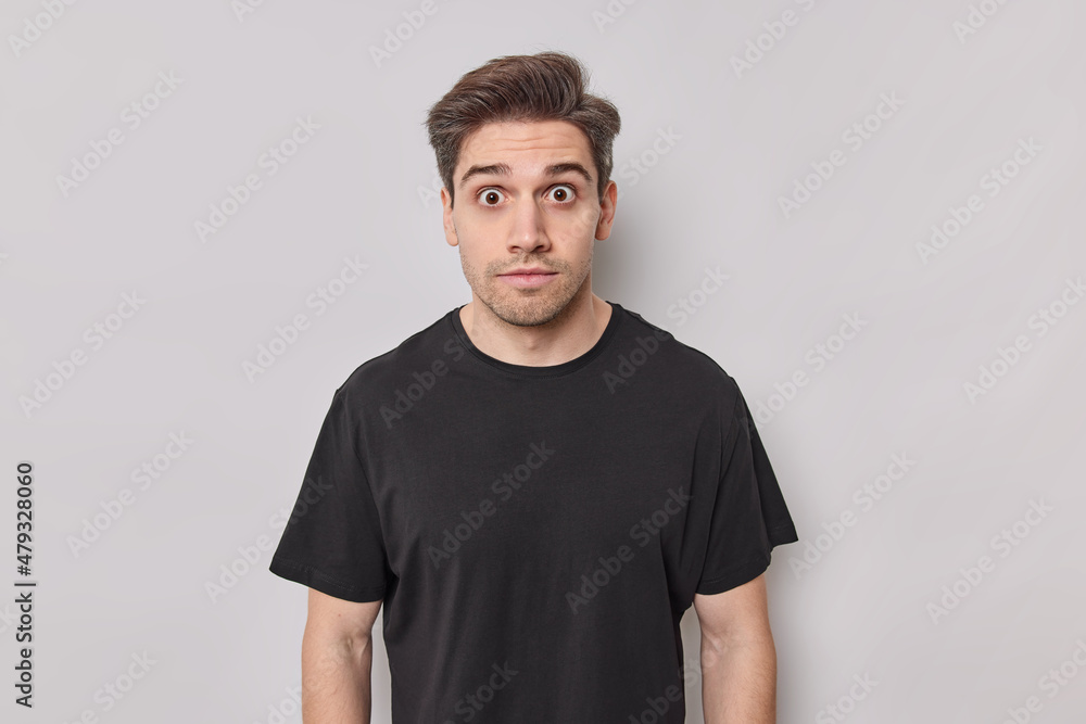 People emotions and reactions concept. Stunned handsome man stares with eyes full of fear dressed in casual black t shirt poses against white background stunned by shocking news stands speechless People emotions and reactions concept. Stunned handsome man stares with eyes full of fear dressed in casual black t shirt poses against white background stunned by shocking news stands speechless
