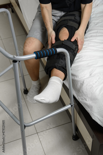 Obraz na płótnie Woman wearing adjustable knee brace after leg fracture sitting on bed trying to get up with walker. Recovery and rehabilitation after surgery and installation of plates. w salonie