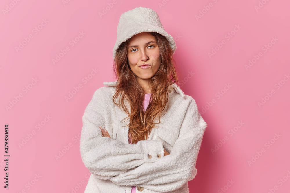 Horizontal shot of self confident woman looks seriously at camera has cheeky expression wears fur hat and coat isolated over pink background. Fashionable good looking female model in outerwear Horizontal shot of self confident woman looks seriously at camera has cheeky expression wears fur hat and coat isolated over pink background. Fashionable good looking female model in outerwear