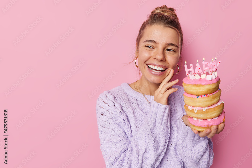 Horizontal shot of attractive woman with combed hair keeps hand on cheek holds pile of doughnuts with burning candles wears knitted sweater isolated over pink background. Celebration concept Horizontal shot of attractive woman with combed hair keeps hand on cheek holds pile of doughnuts with burning candles wears knitted sweater isolated over pink background. Celebration concept