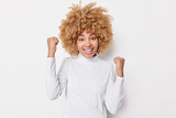 Positive lucky young woman with curly bushy hair celebrates good news clenhches fists from joy receives good news dressed in casual turtleneck isolated over white background. Victory celebration