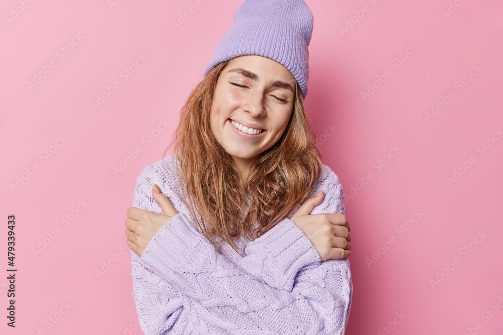 Tender delicate smiling young woman embraces herself with love enjoys comfort in knitted sweater keeps eyes closed wears hat poses against pink background has nice romantic memories feels soft Tender delicate smiling young woman embraces herself with love enjoys comfort in knitted sweater keeps eyes closed wears hat poses against pink background has nice romantic memories feels soft