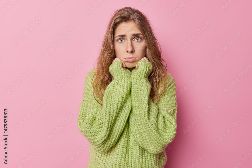 Sad young European woman with long hair has innocent pleading expression keeps hands under chin purses lips wears knitted green sweater isolated over pink background complains about something Sad young European woman with long hair has innocent pleading expression keeps hands under chin purses lips wears knitted green sweater isolated over pink background complains about something