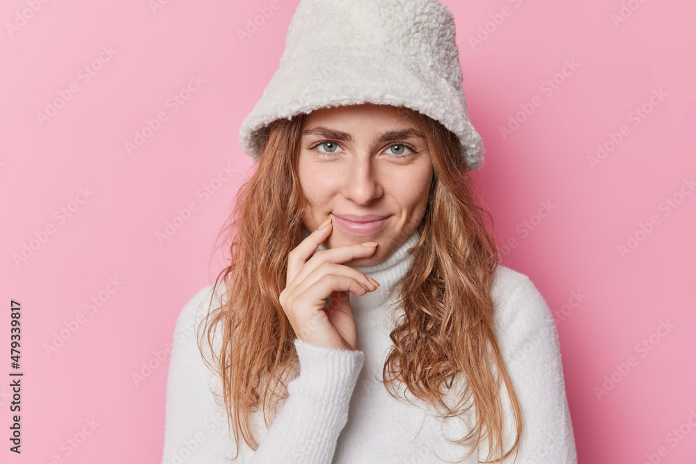 Portrait of good looking young woman looks directly at camera with satisfied expression smiles pleasantly wears white panama and turtleneck isolated over pink background listens positive news Portrait of good looking young woman looks directly at camera with satisfied expression smiles pleasantly wears white panama and turtleneck isolated over pink background listens positive news