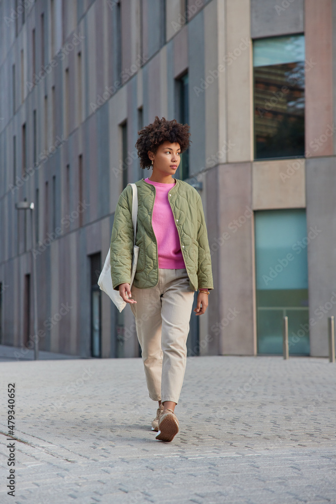 Full length shot of curly haired woman wears jacket trousers and sneakers carries fabric bag strolls outdoors in city centre near modern building reaches necessary destination. Urban lifestyle
