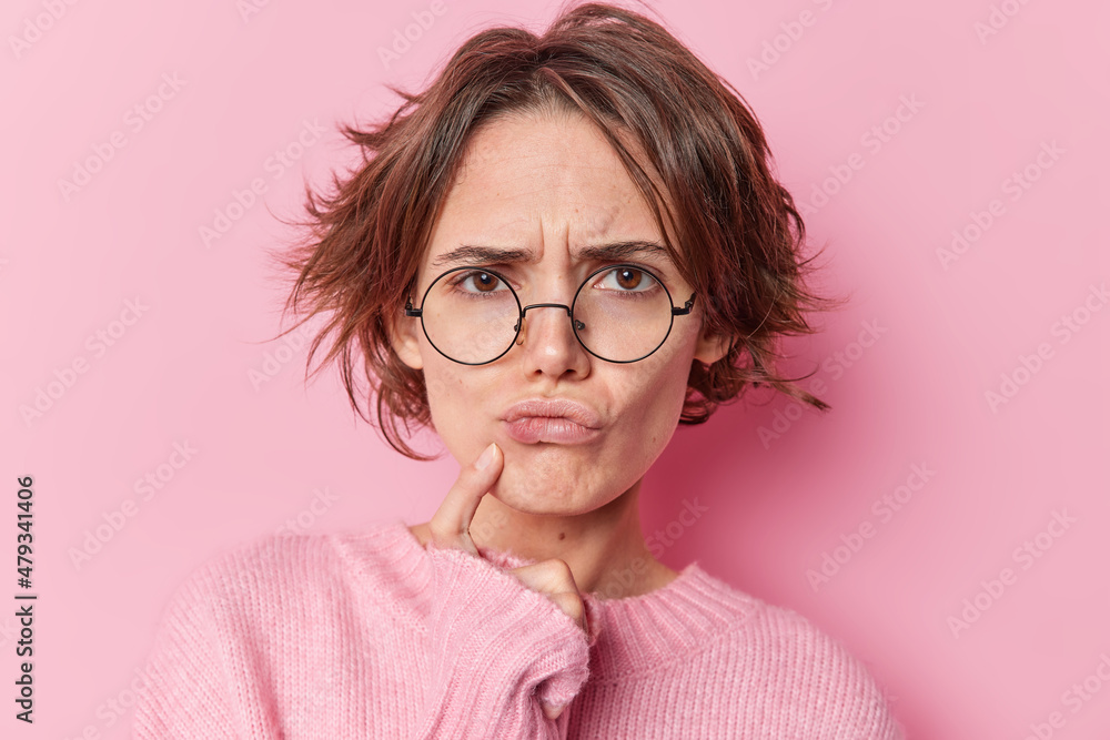 Serious European woman tries to find decision thinks hard keeps finger near lips feels displeased has gloomy expression wears round spectacles long sleeved jumper isolated over pink background - obrazy, fototapety, plakaty 