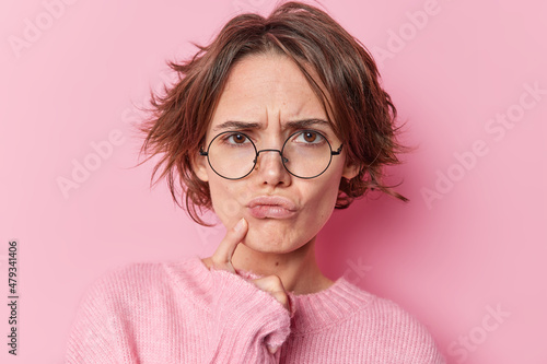 Serious European woman tries to find decision thinks hard keeps finger near lips feels displeased has gloomy expression wears round spectacles long sleeved jumper isolated over pink background