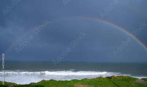 "La corniche de Urrugne" coast of the French Basque country, two rainbows rise parallel to the dark and embroiled sea, a young woman in the distance, on the rocks of the coast, looks at them, below th