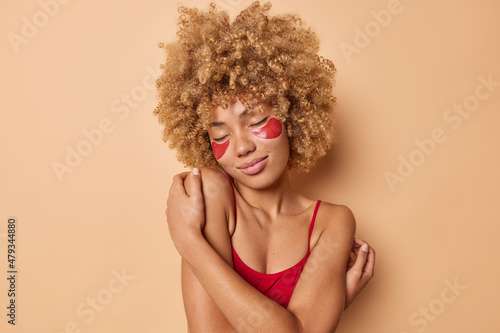 Pleased curly haired young woman expresses self love embraces herserlf touches shoulders gently stands with closed eyes applies red hydrogel patches isolatedover beige background. Beauty concept
