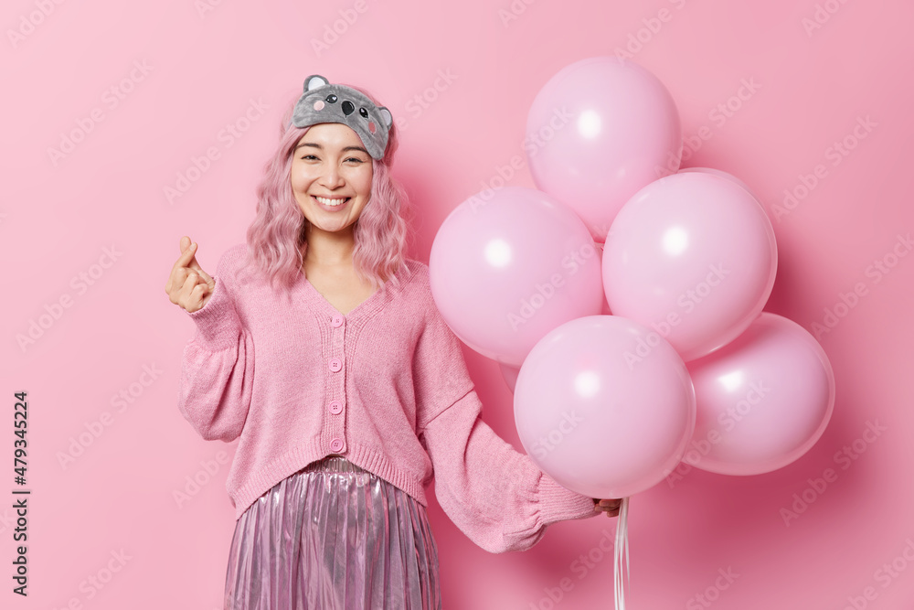Fototapeta premium Happy positive Asian woman makes korean like sign shapes mini heart expresses love holds bunch of balloons wears sleepmask jumper and skirt celebratess birthday isolated over pink background