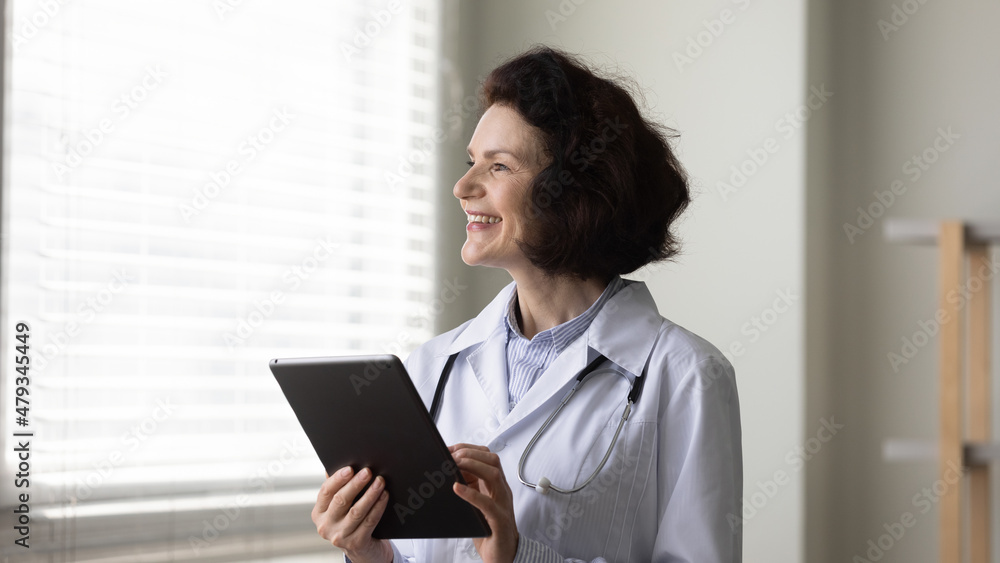 Dreamy happy old senior 50s female GP doctor physician holding digital computer tablet in hands, looking in distance, thinking of professional career opportunities, medicare and modern tech concept. Dreamy happy old senior 50s female GP doctor physician holding digital computer tablet in hands, looking in distance, thinking of professional career opportunities, medicare and modern tech concept.