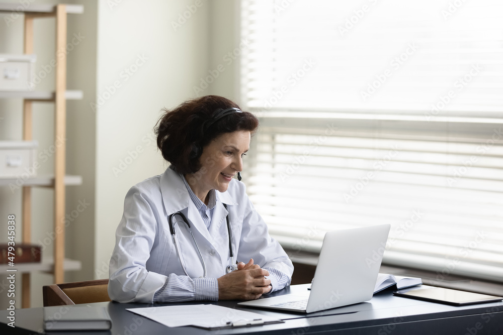 Happy older mature female doctor therapist physician in headset with mic looking at laptop screen, consulting patient remotely by distant web camera video call meeting in clinic, telemedicine concept. Happy older mature female doctor therapist physician in headset with mic looking at laptop screen, consulting patient remotely by distant web camera video call meeting in clinic, telemedicine concept.