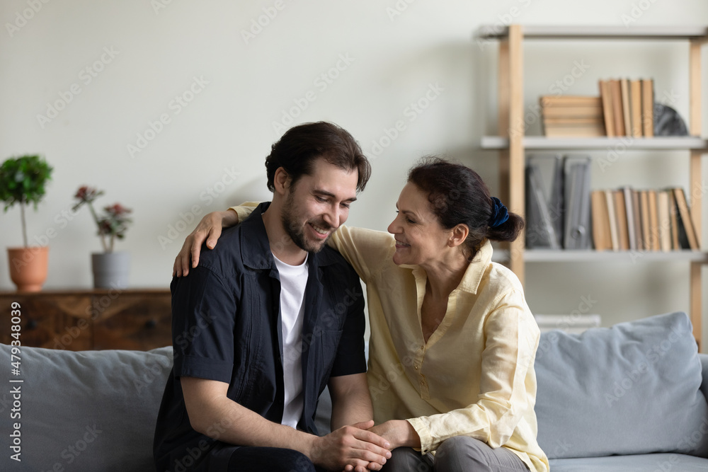 Happy old retired woman cuddling shoulder of grown smiling son, gossiping or enjoying trusted conversation, sitting on cozy couch together at home, spending weekend time together, relations concept. Happy old retired woman cuddling shoulder of grown smiling son, gossiping or enjoying trusted conversation, sitting on cozy couch together at home, spending weekend time together, relations concept.