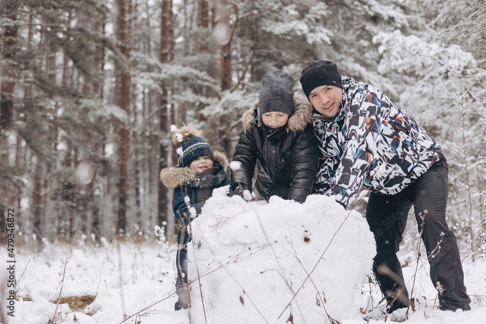 Happy father and sons rolling big snowball together for snowman in snowy winter forest. Man Joyful teenager and little boy playing and having fun on family walking. Wintertime activity outdoors.
