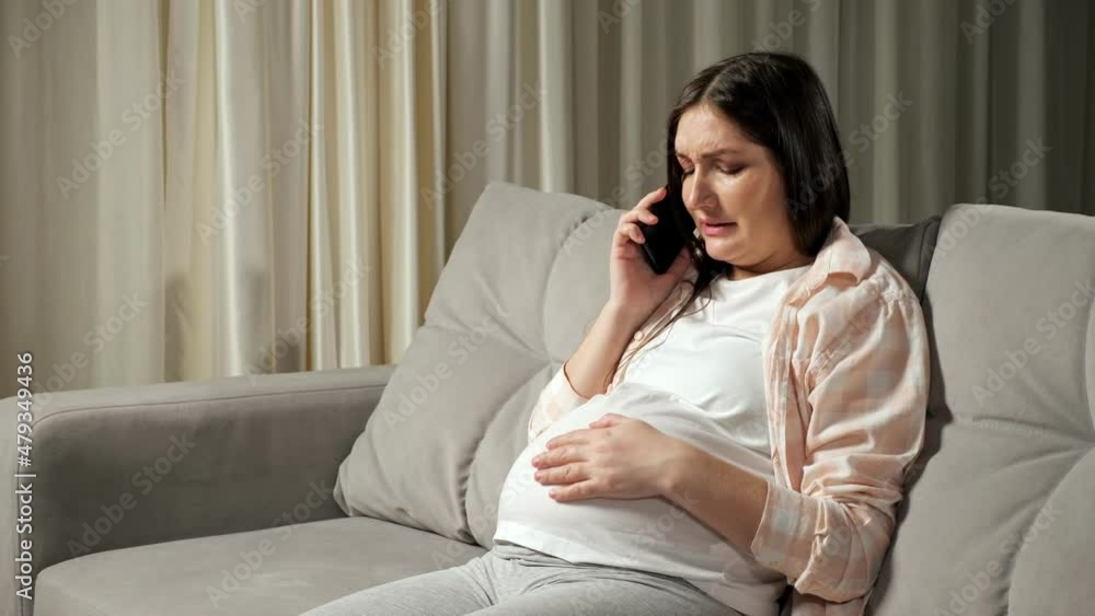 Pregnant woman with long brunette hair feels contractions and calls on husband for help breathing heavily and stroking belly in pain on sofa. Pregnant woman with long brunette hair feels contractions and calls on husband for help breathing heavily and stroking belly in pain on sofa.