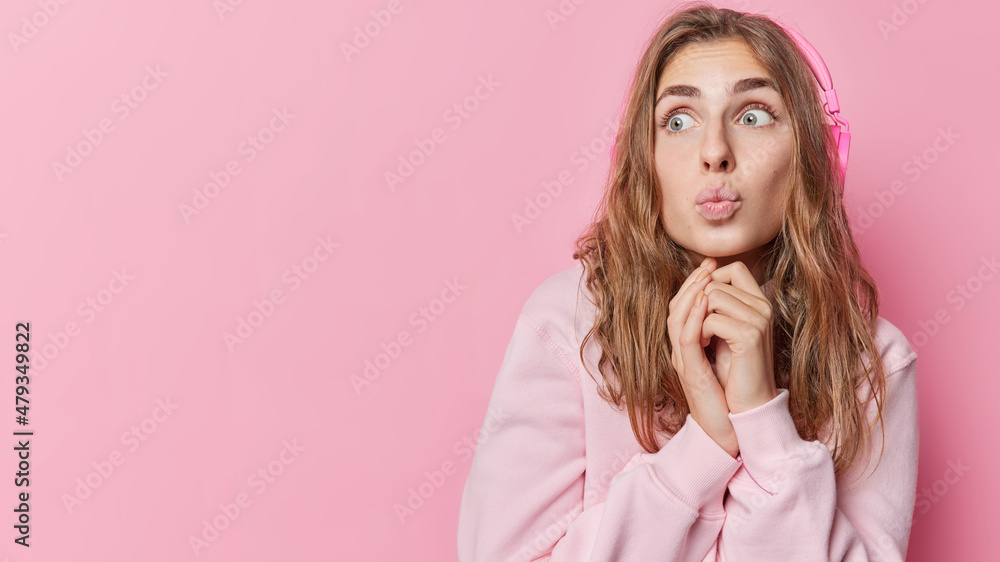 Horizontal shot of impressed young woman keeps lips rounded looks away surprisingly listens favorite music via headphones wears long sleeved jumper isolated over pink background empty space. Horizontal shot of impressed young woman keeps lips rounded looks away surprisingly listens favorite music via headphones wears long sleeved jumper isolated over pink background empty space.