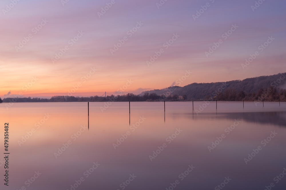 Early winter morning during a vibrant sunrise full of colors and with a veil of fog over the lake. 
The fishing poles create nice reflections at the Pietersplas in Maastricht