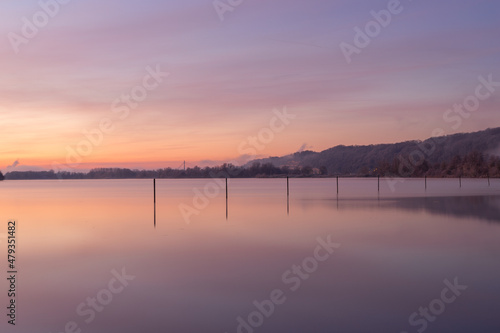 Early winter morning during a vibrant sunrise full of colors and with a veil of fog over the lake. The fishing poles create nice reflections at the Pietersplas in Maastricht