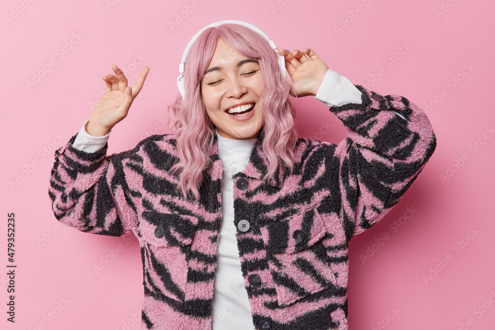 Carefree positive Asian woman with dyed hair moves with rhythm of music wears wireless headphones on ears dressed in winter coat keeps eyes closed smiles broadly isolated over pink background Carefree positive Asian woman with dyed hair moves with rhythm of music wears wireless headphones on ears dressed in winter coat keeps eyes closed smiles broadly isolated over pink background