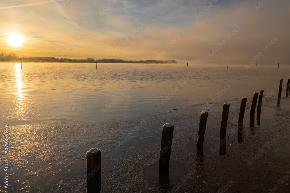 A sunrise seen from the shore of a lake during winter time. The rising sun reflects in the thin layer of ice on the water between the poles of the edge of the lake.