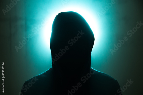 a man in a hood without a face, a light shines behind his back, a photo indoors