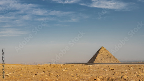 The ancient Egyptian pyramid of Mykerin on the background of the blue sky. Silhouettes of modern high-rise buildings of Cairo are visible in the distance. In the foreground  yellow desert sand  rocks.