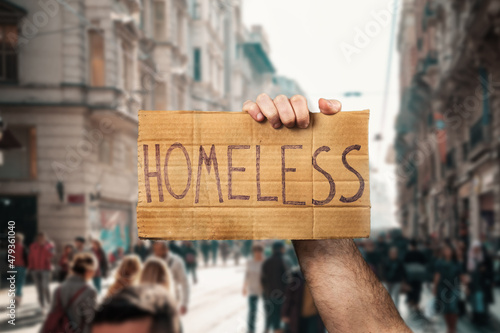 A man hold a cardboard sign with the inscription HOMELESS. Defocused background with street and people. Hands close-up. The concept of helping vagabonds
