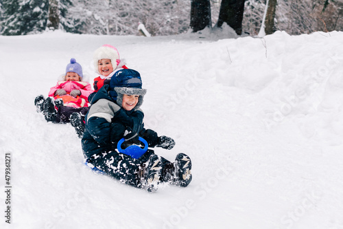 Little caucasian kids in warm clothing laughing looking at camera and toboganning on snow covered hill. Full lengh horizontal shot. Selective focus. Happy childhood and active wintertime concept.