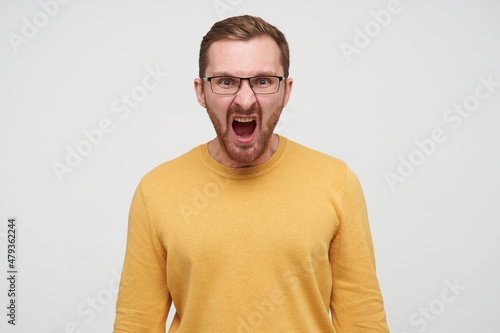 Studio portrait of young bearded male student wears yellow sweater screaming into camera with angry facial expression, isolated over white background © timtimphoto