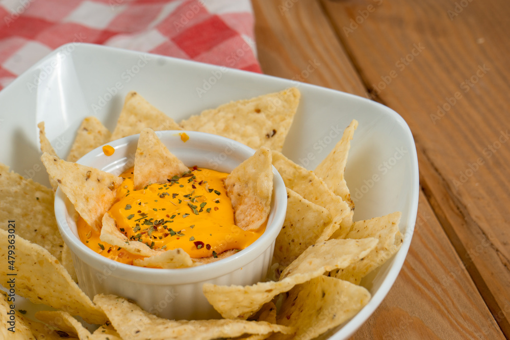 Nachos dipped in cheddar on a bowl on a wooden table. Under the dish there is a red and white kitchen cloth and the background is grey. Vertical image