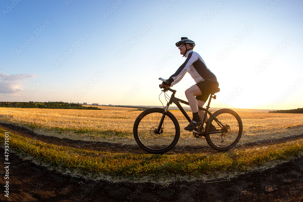 The cyclist rides a bike on the road near the field against the backdrop of the setting sun. Outdoor sports. Healthy lifestyle.