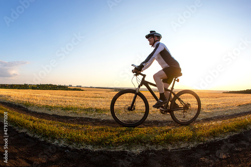 The cyclist rides a bike on the road near the field against the backdrop of the setting sun. Outdoor sports. Healthy lifestyle. © photosaint