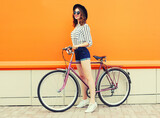 Summer colorful portrait of happy smiling young woman with bicycle in the city on orange background