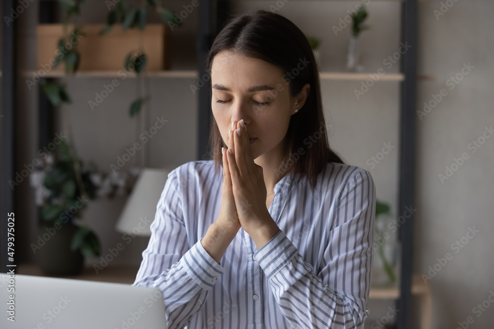 Religious millennial business woman praying at workplace. Female prayer, employee, worker asking, worshipping God, begging for help, healing, good luck, working at desk with laptop. Religion concept Religious millennial business woman praying at workplace. Female prayer, employee, worker asking, worshipping God, begging for help, healing, good luck, working at desk with laptop. Religion concept
