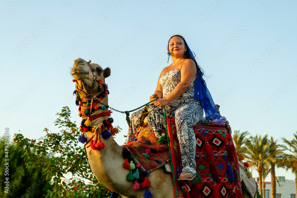 A Jewish woman in a cape on her head rides a camel on a city street against the backdrop of palm trees.