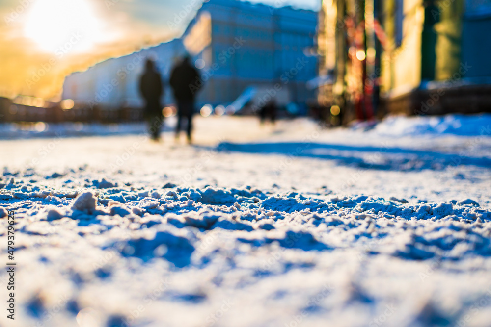 Winter in the city. A couple are walking down the street. Snow cover. Historical center of the city. Sunset. Focus on snow. Close up view from the level of the road. Winter in the city. A couple are walking down the street. Snow cover. Historical center of the city. Sunset. Focus on snow. Close up view from the level of the road.