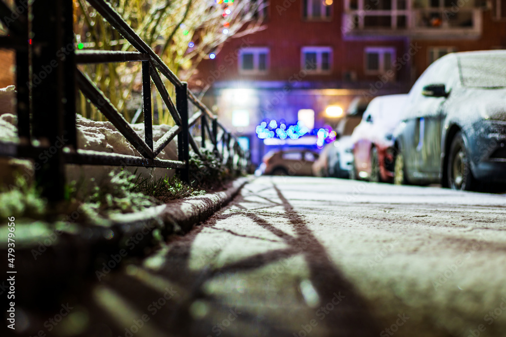 Winter in the night city. Sidewalk in the yard. Residential area. Snowy road. A row of parked cars. Working lights. Snowfall. Juicy colors. Focus on snow. Close up view from sidewalk level.
