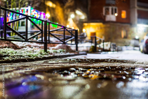 Winter in the night city. Sidewalk in the yard. Residential area. Working lights. Snowfall. Juicy colors. Christmas decorations. Focus on the tiled sidewalk. Close up view from sidewalk level.