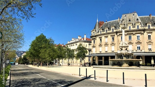 Valence Drôme France Boulevard avec de beaux immeubles haussmanniens et sa fontaine monumentale sur un ciel bleu ensoleillé.