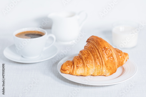 Croissant in bright white environment, with cup of coffee and milk and other croissant in blurry background. Simple and elegant breakfast setting and scene.