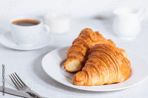 Croissant in bright white environment, with cup of coffee and milk and other croissant in blurry background. Simple and elegant breakfast setting and scene.