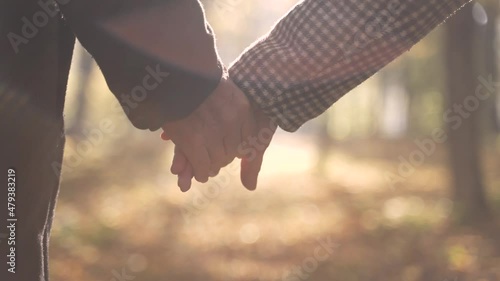 Close-up of joining wrinkled hands of aged loving husband and wife during walk on road to heaven to bright sunlight. Happy old pair taking each other's hand while walking towards sun in autumn park