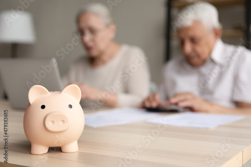 Close up focus on small piggybank on table with blurred middle aged old family couple doing financial paperwork, managing household budget or planning investment on background, retirement lifestyle.
