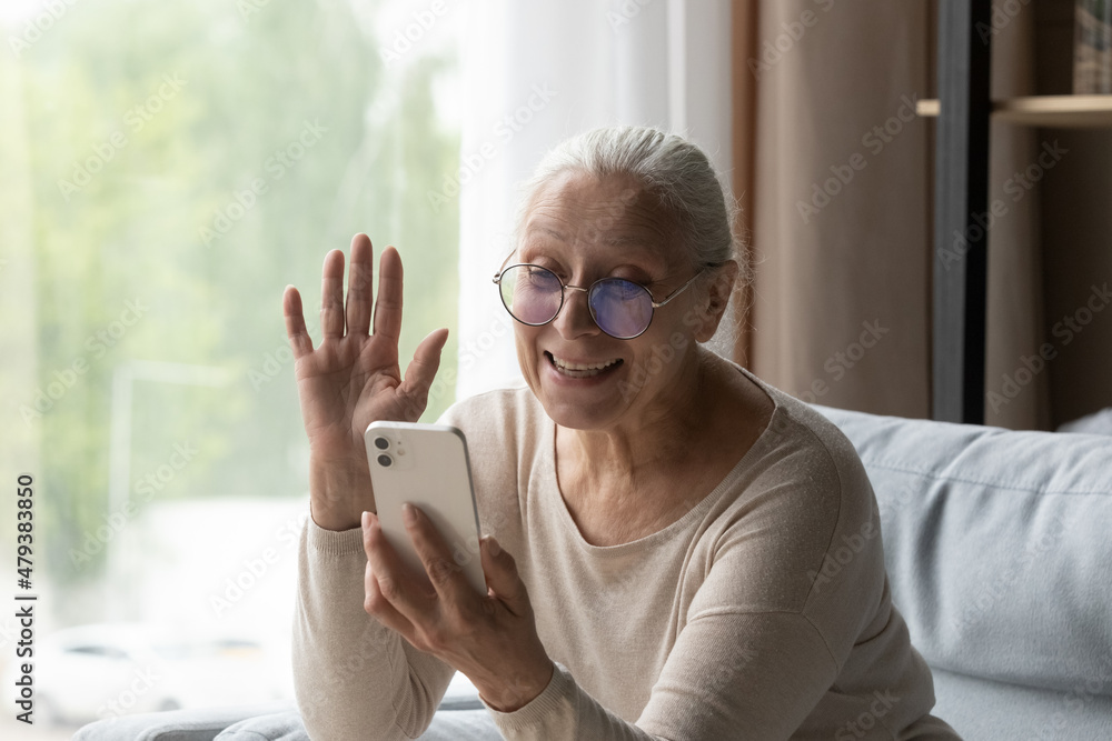 Happy old mature retired grandmother in eyeglasses waving hand, looking at smartphone web camera, starting video call distant conversation with grownup children or friends, chatting online at home.