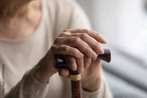 Close up focus on folded wrinkled female hands on wooden cane. Cropped thoughtful elderly senior grandmother having walking disability  using stick indoors  old retired people lifestyle concept.