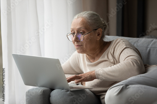 Addicted to modern technology happy middle aged woman in eyeglasses using laptop software applications, enjoying web surfing information, communicating in social network, playing games or shopping.