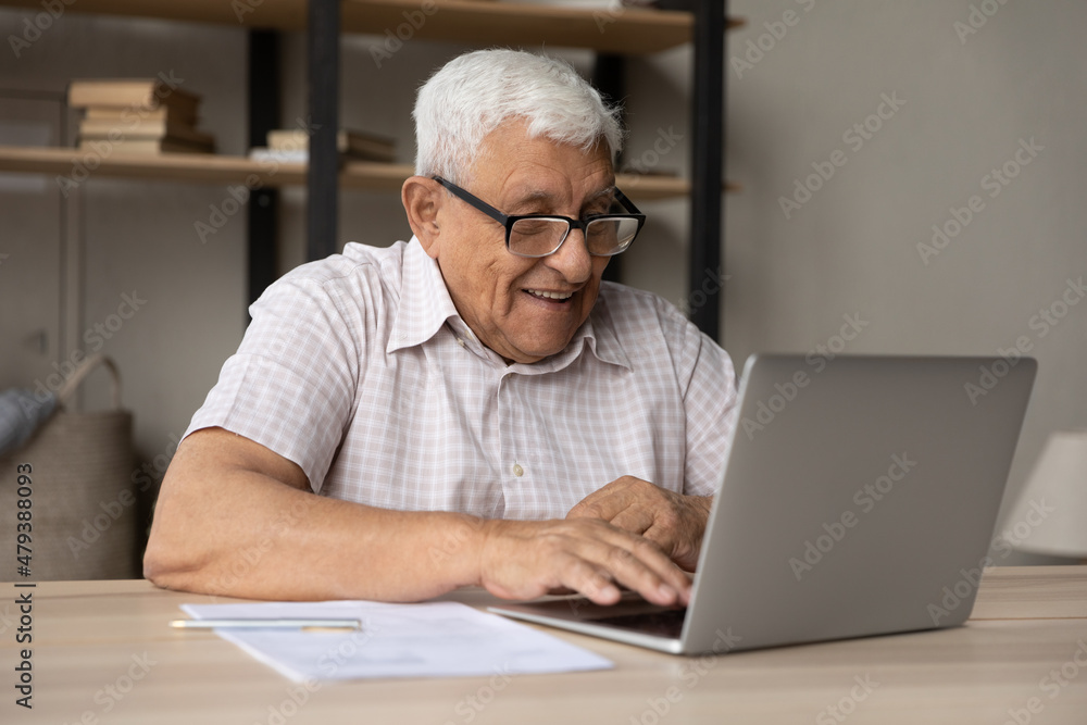 Smiling mature elderly retired man in eyeglasses working on computer, sitting at table at home or office, enjoying communicating in social network, checking email, web surfing or shopping online. Smiling mature elderly retired man in eyeglasses working on computer, sitting at table at home or office, enjoying communicating in social network, checking email, web surfing or shopping online.